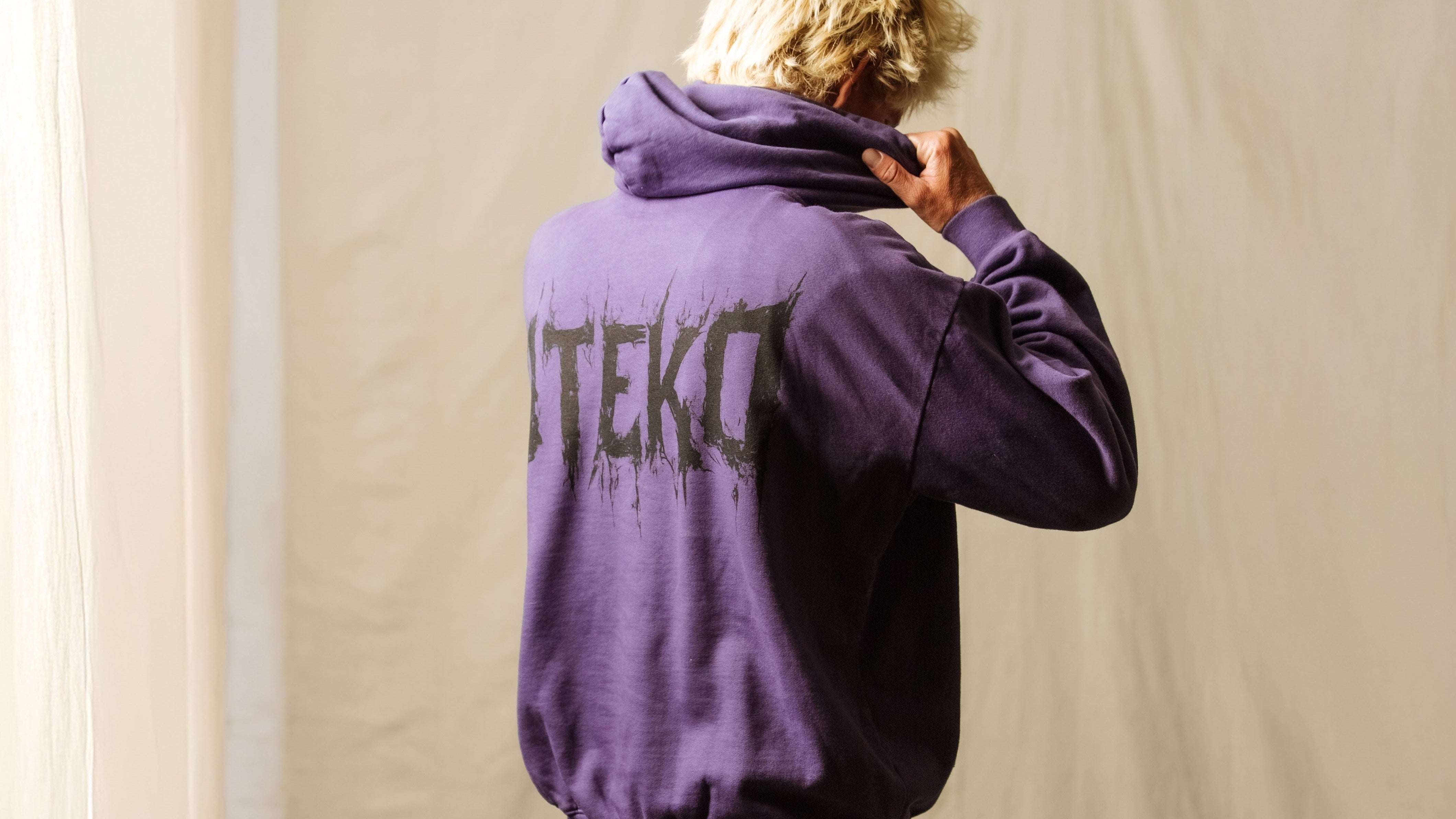Steko brand lookbook photograph, a model in a faded purple Steko hoodie with the brand's scratched logo across the back